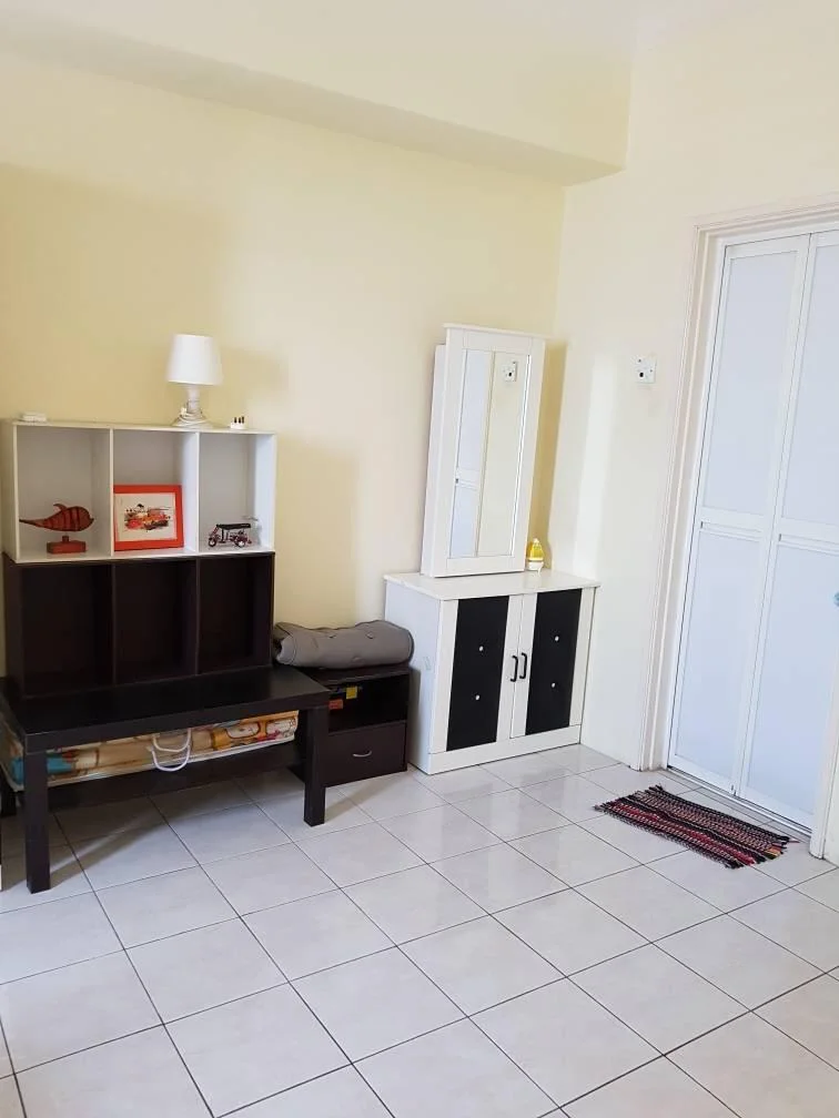 Room features: white painted walls, glossy ceramic tile flooring, black and white storage cabinet, open shelving unit with decorative items, small white lamp on shelf, grey blanket on shelf, door with white frame, colorful doormat; style: modern.